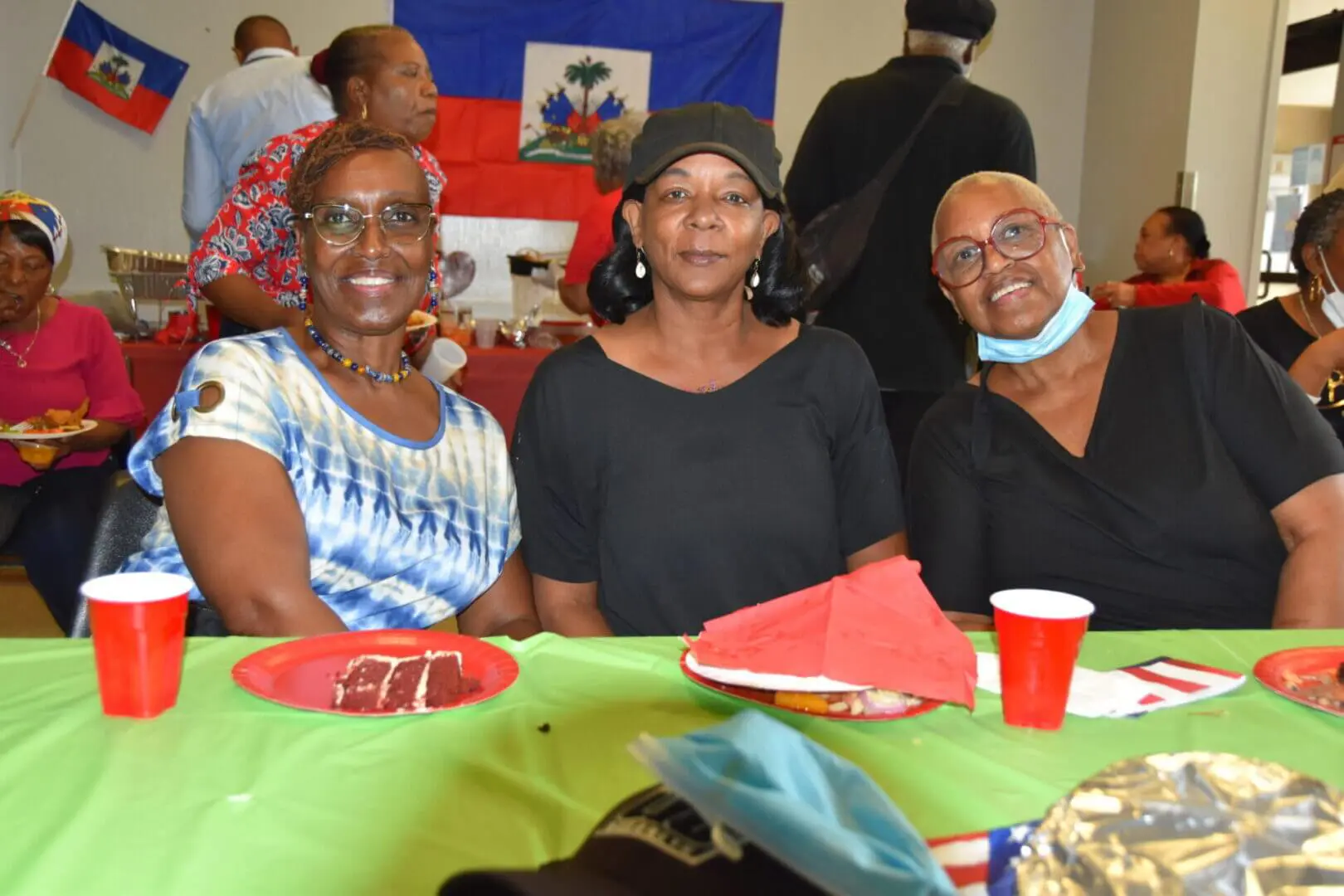 Three people smiling and sitting at a table with watermelon slices and a red cup.