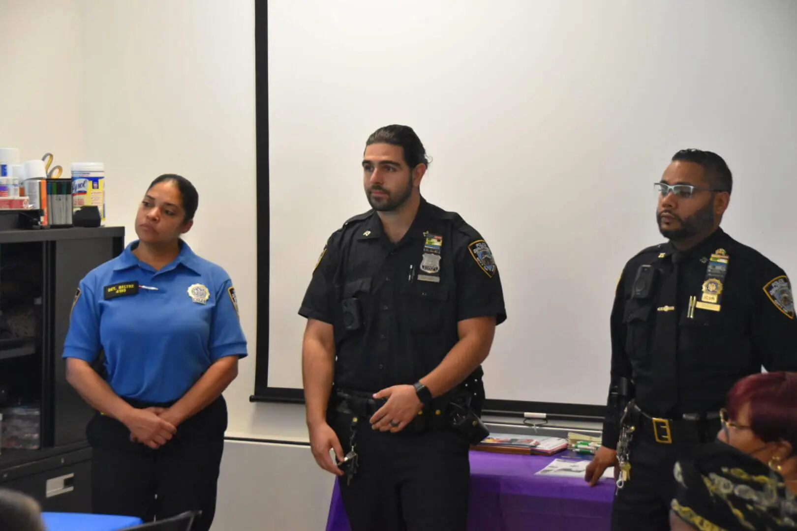 Three security personnel standing indoors near a table with purple cloth.
