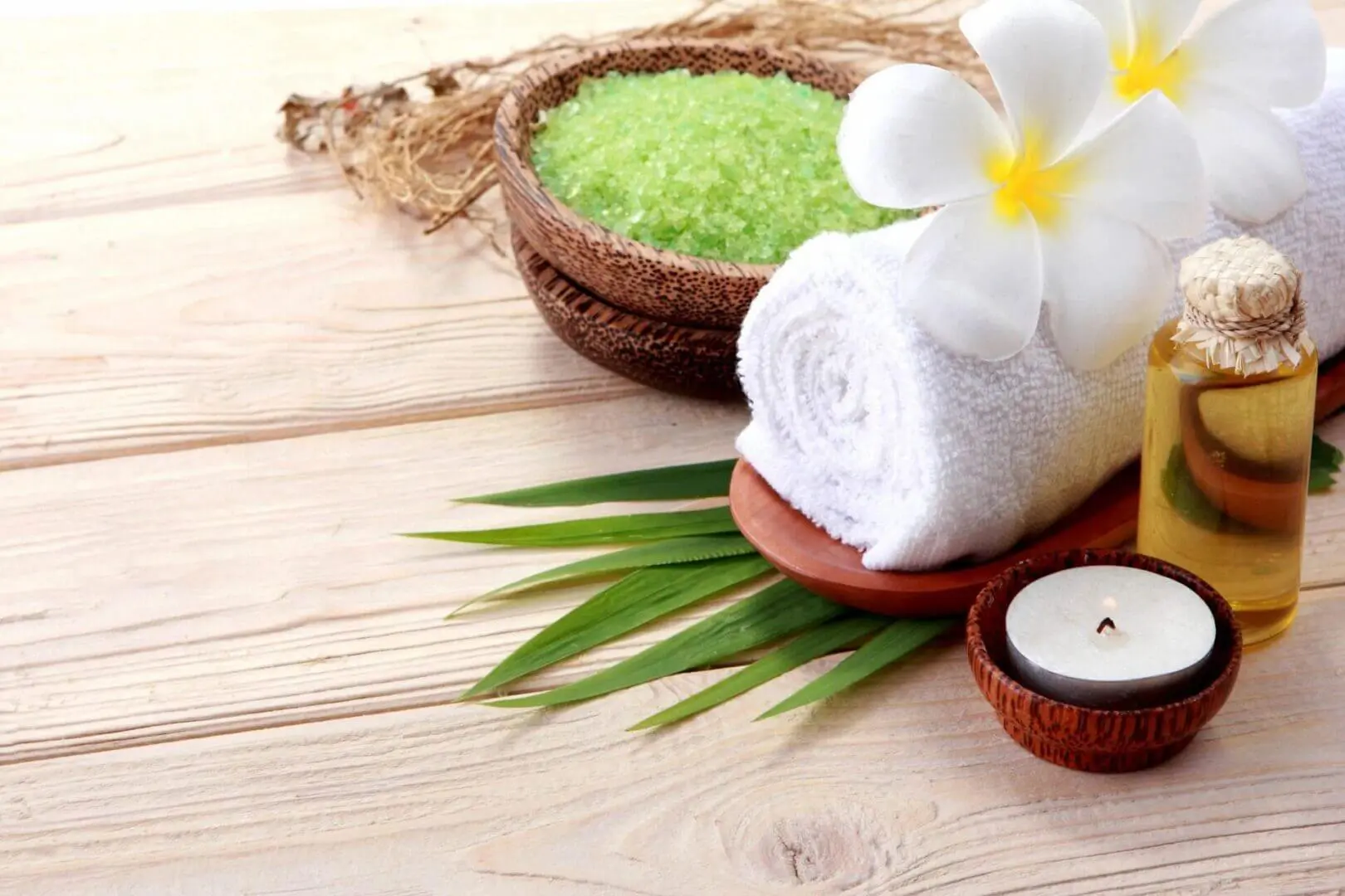 Spa essentials including green bath salts, white towel, and candle on wooden surface.