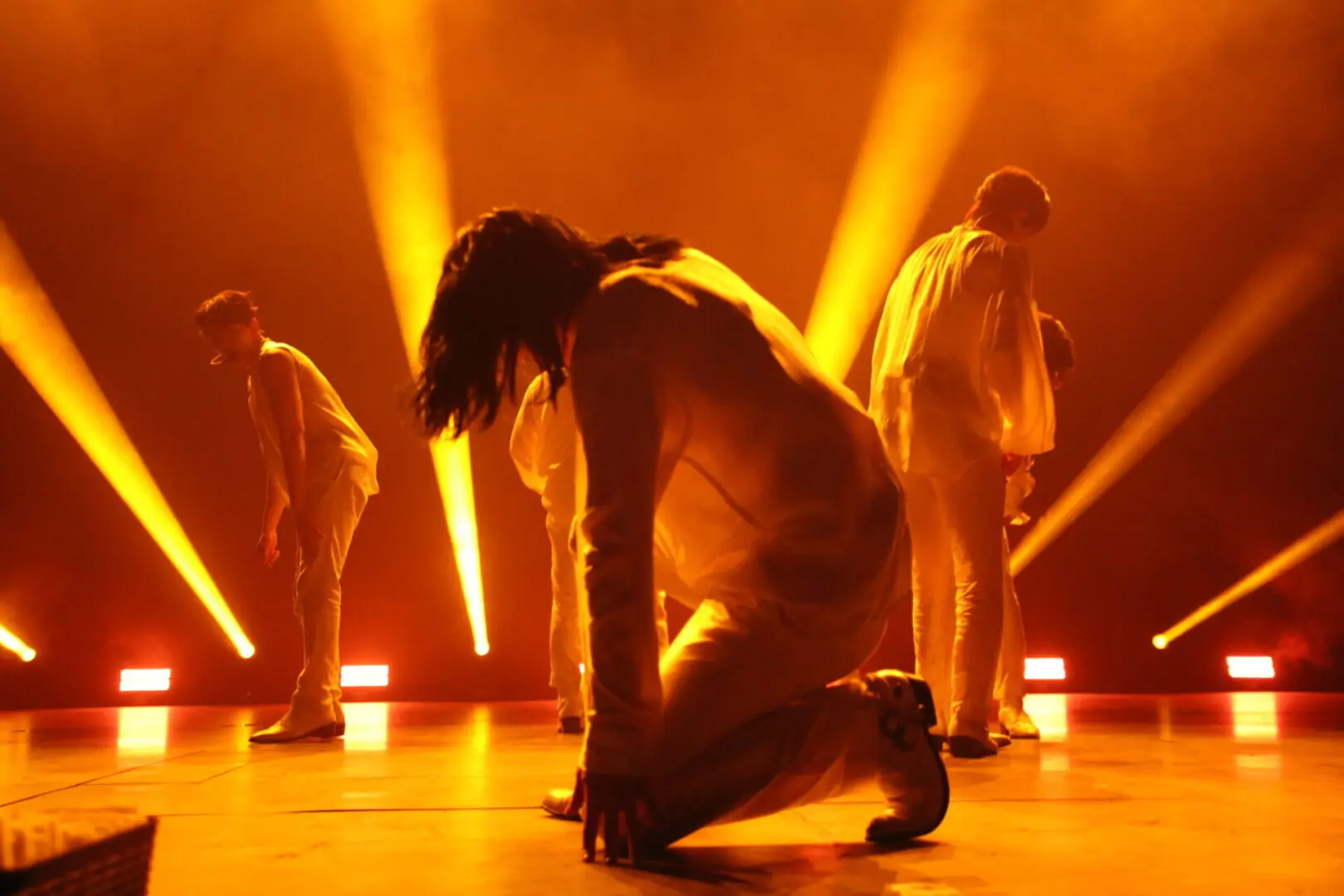 Dancers in white costumes perform under vibrant orange stage lights.