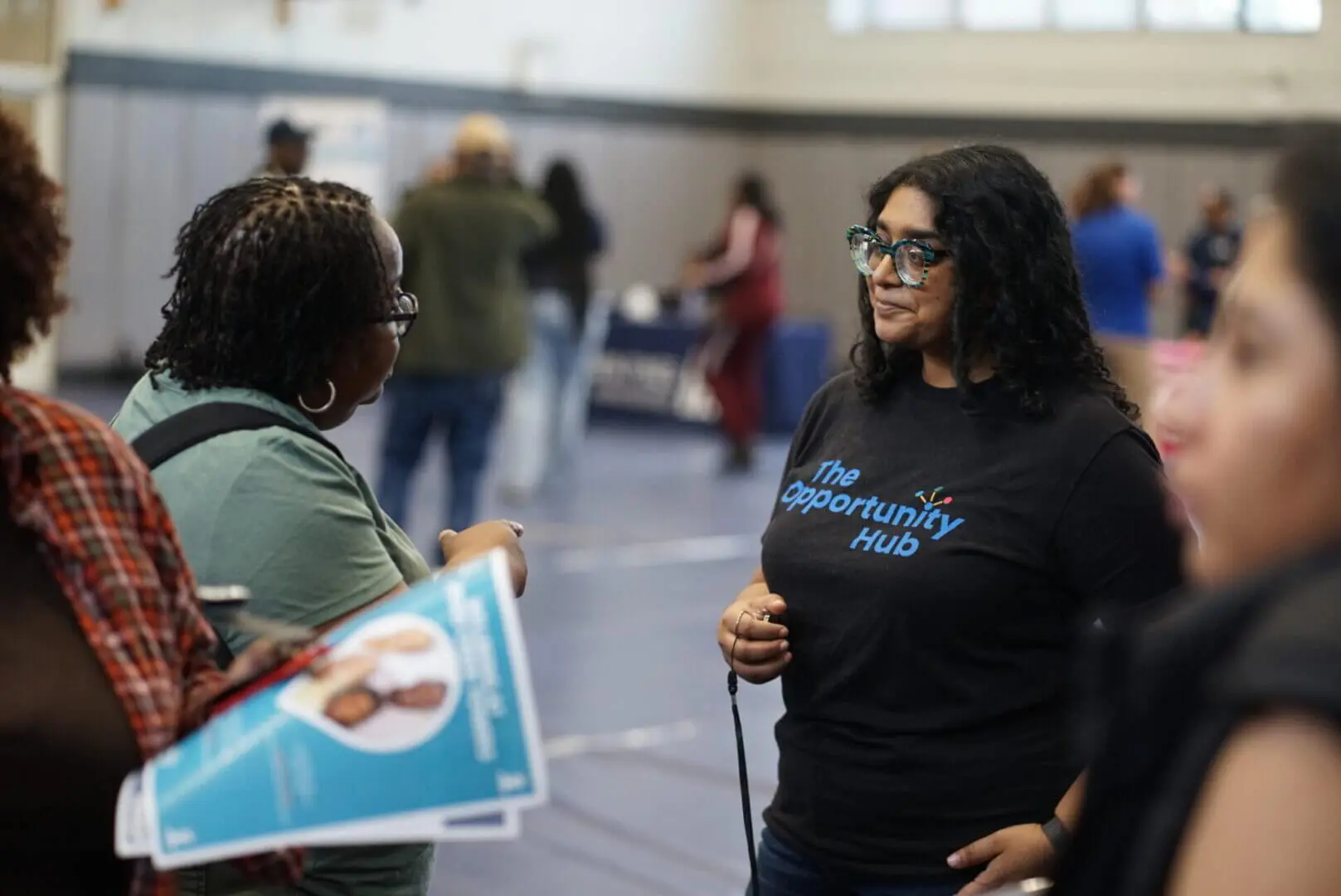 Two women engage in conversation at an indoor event with others in the background.