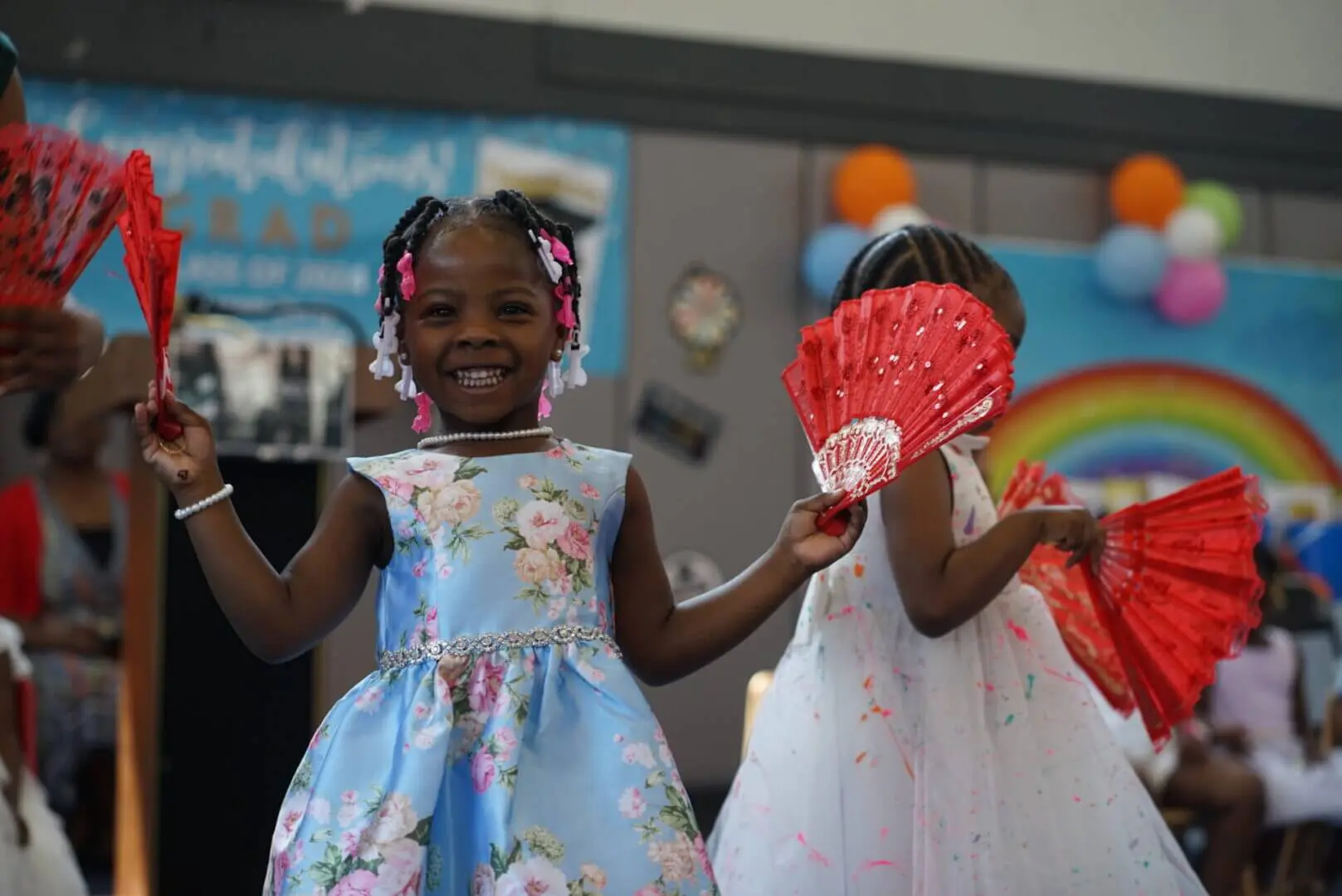 Two young girls happily playing with fans, dressed in colorful dresses.