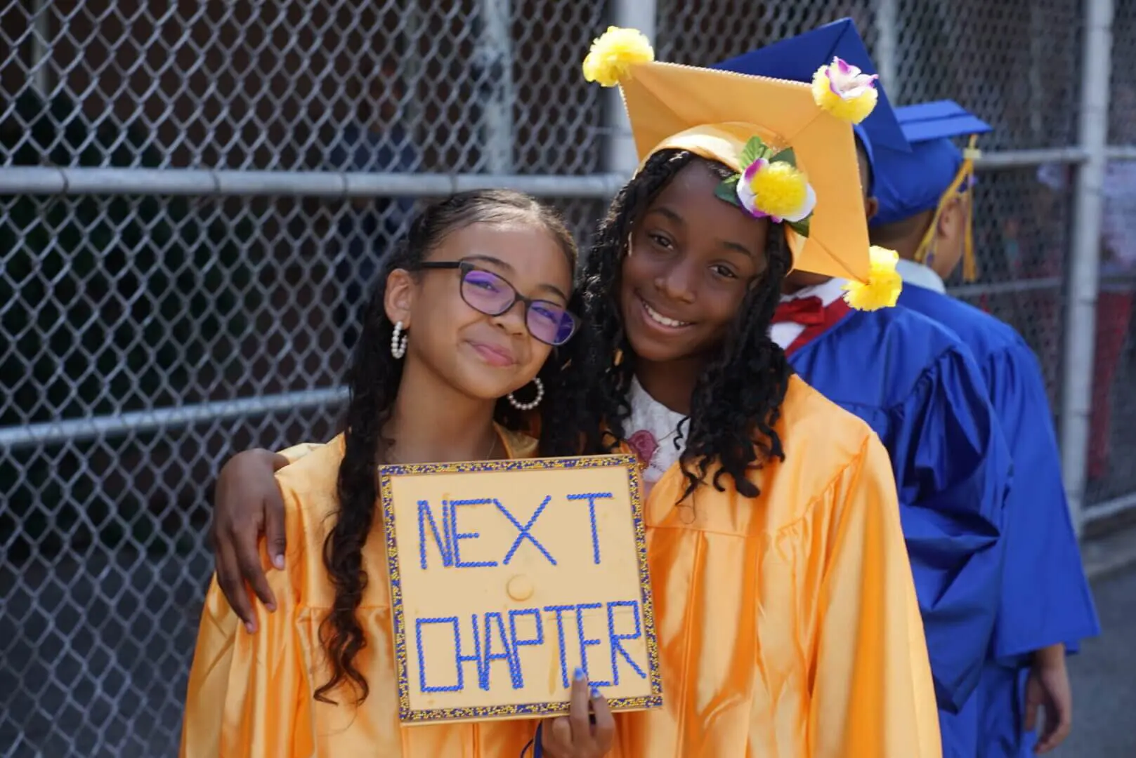Two graduates smiling, one holding a 'Next Chapter' sign.