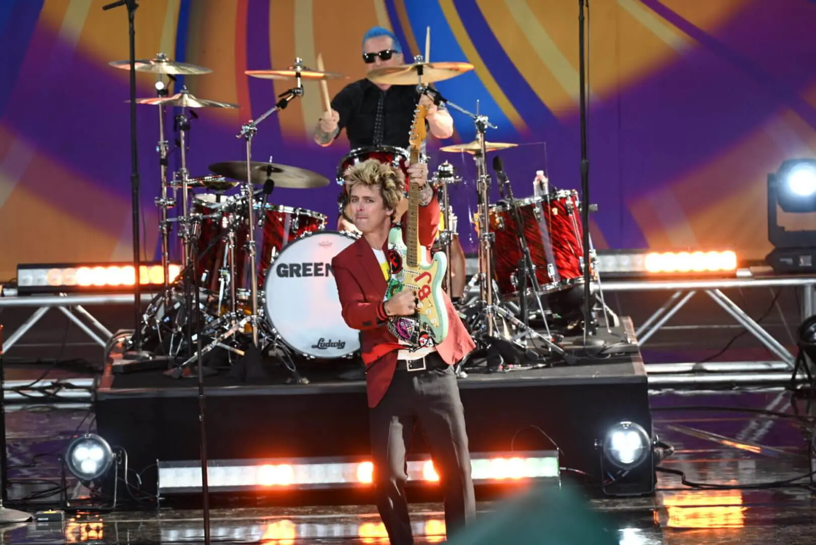 Musician playing guitar on stage with drummer in background.