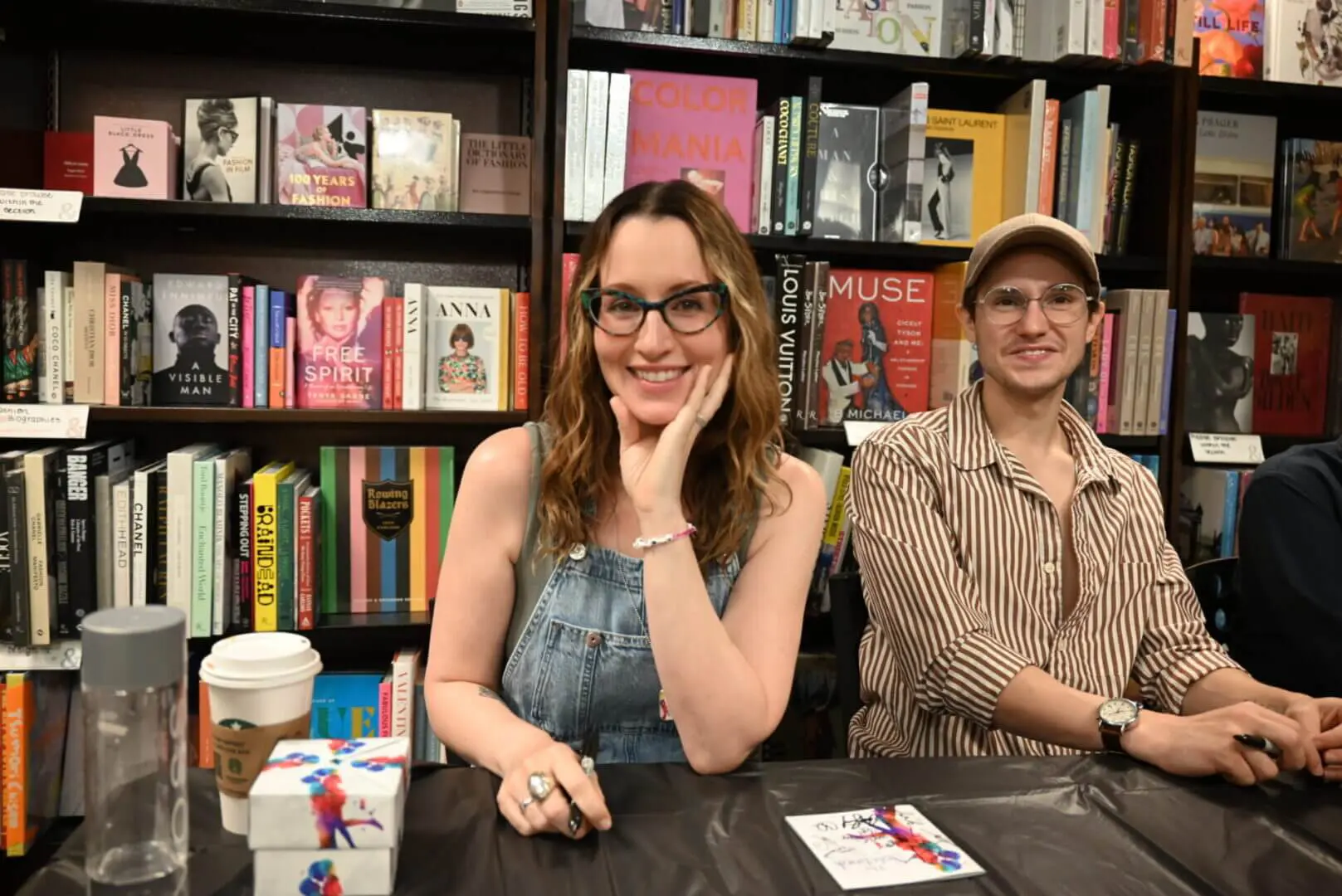 Two people smiling at a book signing event in front of shelves filled with books.