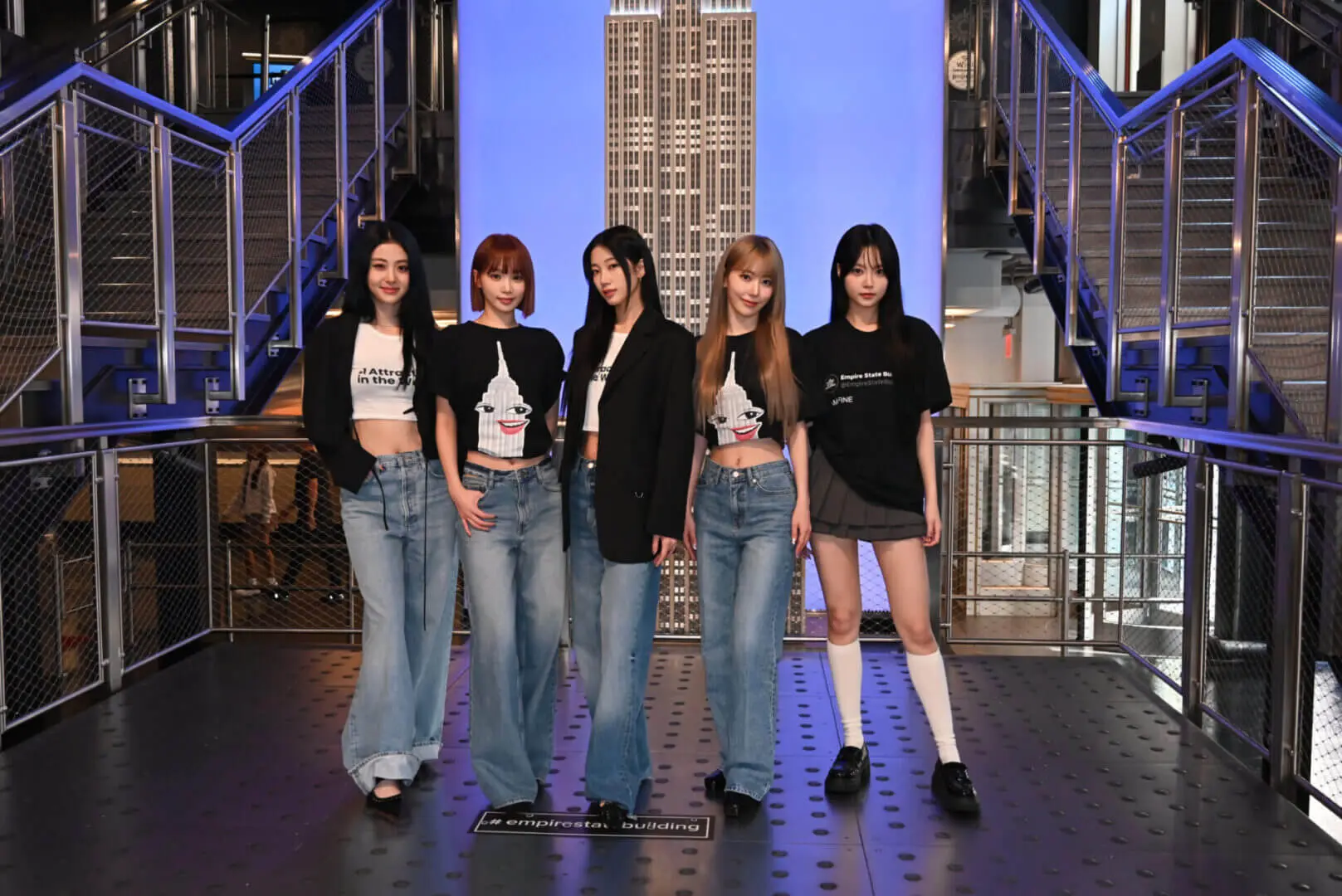 Five young women posing stylishly on a staircase with cityscape backdrop.
