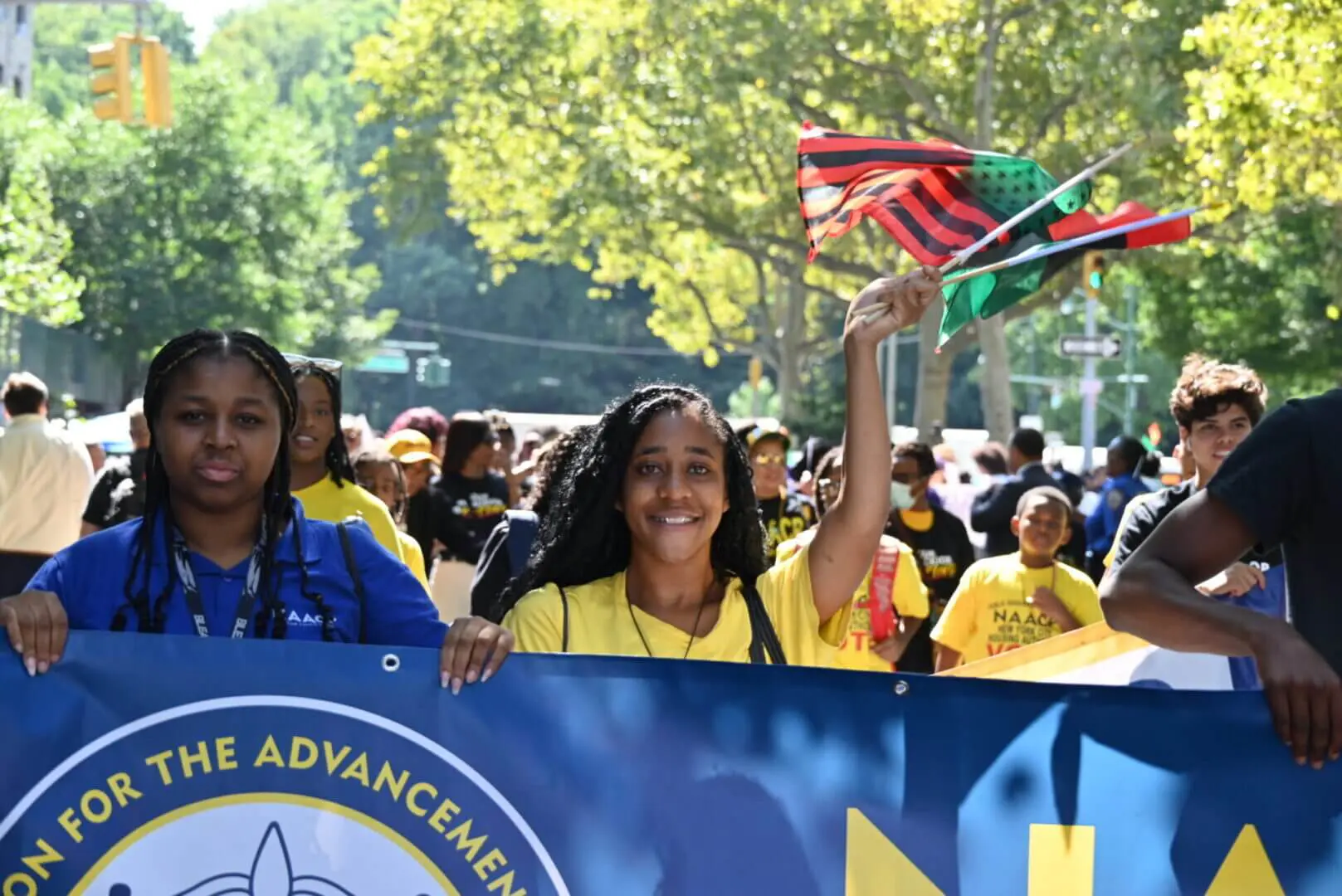 Young women joyfully holding a colorful banner during a sunny outdoor event.