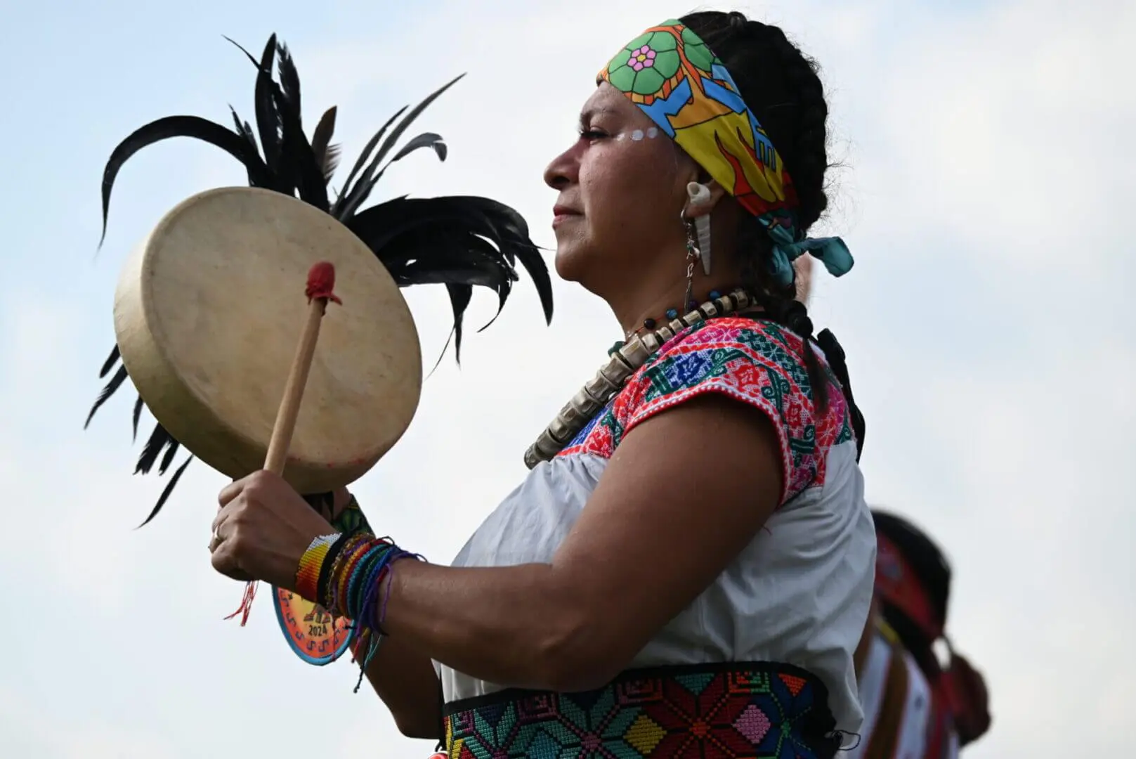 Indigenous woman playing a traditional drum in colorful attire.