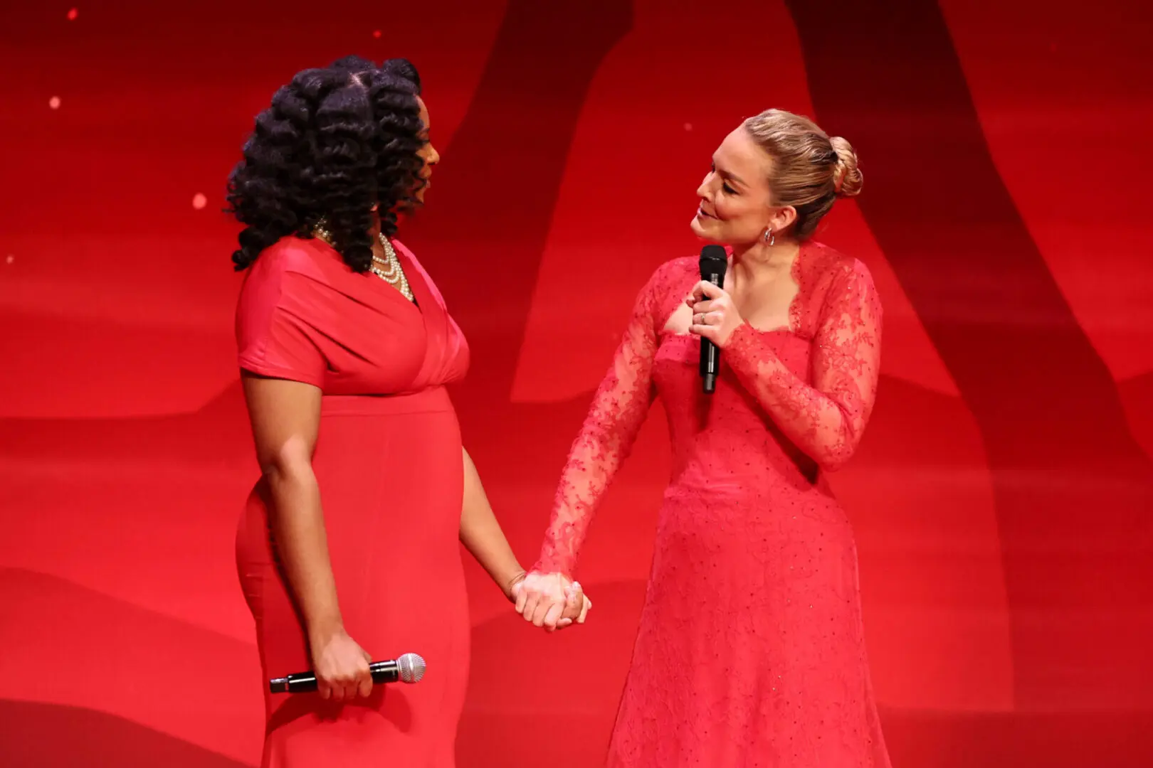 Two women in red dresses holding hands and smiling on stage with microphones.